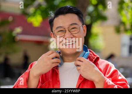 Portrait d'homme d'âge moyen asiatique souriant heureux écoutant de la musique décollant des écouteurs regardant la relaxation de l'appareil photo se sentir satisfait de bonnes nouvelles en plein air. Mec mûr chinois dans la rue de la ville. Horizontal Banque D'Images