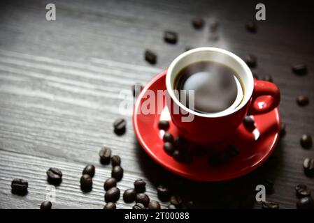 Café de tasse rouge avec vapeur de ruisseau et grain de café sur la table en bois, focalisation sélective sur le bord de la tasse, concept de boisson Banque D'Images