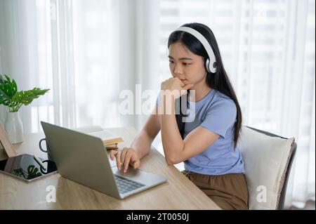 Une jeune femme asiatique concentrée et réfléchie portant des écouteurs, écoutant de la musique tout en travaillant sur son ordinateur portable à une table dans sa chambre. apprentissage en ligne, Banque D'Images