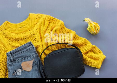 Pose à plat de pull jaune de femme, jeans gris, sac noir et petite citrouille, feuille d'automne sur fond gris. Concept de mode d'automne. Vue de dessus, plat la Banque D'Images