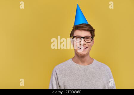 jeune homme joyeux en sweat-shirt blanc et chapeau d'anniversaire souriant à la caméra sur fond jaune Banque D'Images