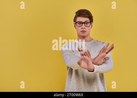 jeune homme triste en sweat-shirt blanc avec des lunettes montrant le geste de la main croisée de déni sur fond jaune Banque D'Images