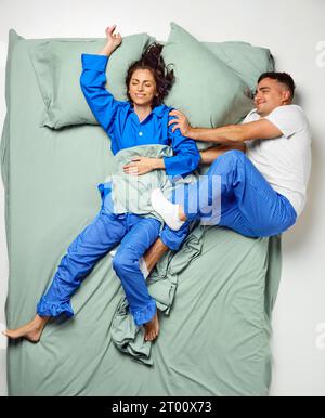 Jeune belle femme couverte de couverture dormant sur le lit et homme couché sur le côté gelant, couple portant en pyjama se relaxant dans le grand lit de menthe. Banque D'Images