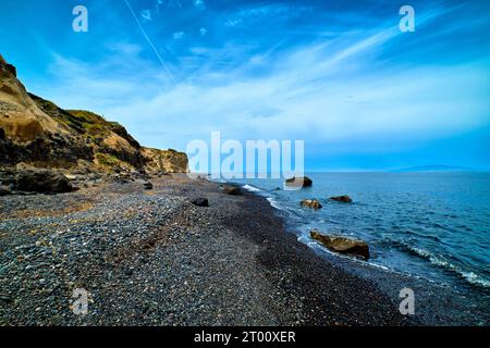 Paysage spectaculaire de la mer bleue ou des vagues de l'océan roulant sur la plage volcanique rocheuse le jour nuageux. Ciel bleu, beaux nuages, galets de lave noirs et sable, falaise patinée. Île de Santorin, Grèce. Banque D'Images