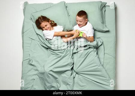 Portrait de vue de dessus de la petite fille et le garçon couché sur le dos recouvert d'une couverture ne peut pas séparer le téléphone. Adorable enfant, enfant dans un grand lit de menthe. Banque D'Images