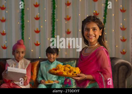 Fille joyeuse debout avec une assiette de fleurs à l'occasion de Diwali et ses amis assis derrière Banque D'Images