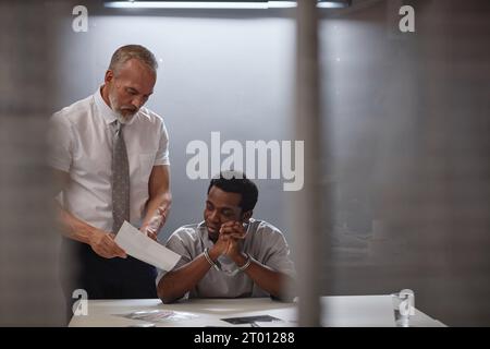 Portrait du jeune homme noir dans la salle d'interrogatoire avec détective senior montrant des photos de preuves, espace de copie Banque D'Images