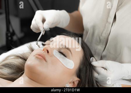 Une esthéticienne regarde dans un mini miroir et regarde les cils nouvellement étendus d'une jeune femme dans un salon de beauté. Le visage de la femme en procédure esthétique Banque D'Images