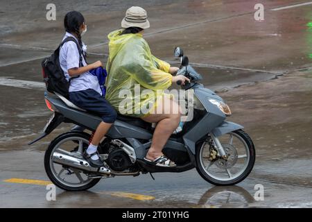 Une femme conduisant une moto avec une fille en uniforme scolaire dans une rue pluvieuse, Thaïlande Banque D'Images