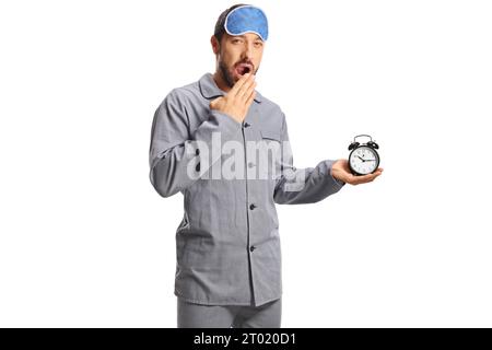 Jeune homme en pyjama avec un masque de sommeil tenant un réveil et bâillant isolé sur fond blanc Banque D'Images