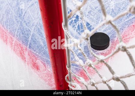 Regarder à travers le filet de but vers un palet de hockey sur glace noir labourant à travers la neige et s'arrêtant sur le bord de la ligne de but du pli de but. Banque D'Images