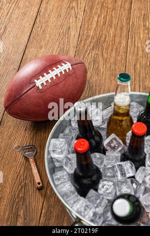 Regardant vers le bas sur un ballon de football américain assis à côté d'une baignoire galvanisée avec des bouteilles de bière assorties, assis sur une table en bois avec un ouvre-bouteille. Banque D'Images