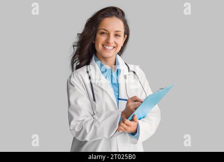 Portrait de jeune femme médecin souriante en uniforme tenant le presse-papiers, regardant et souriant à la caméra, fond gris Banque D'Images