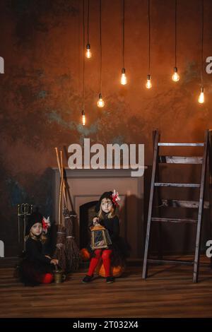 Les filles en costumes de sorcières sont assises près de la cheminée sur des citrouilles, l'une tient une lanterne, l'autre fait une potion. Halloween. Cadre vertical. Banque D'Images