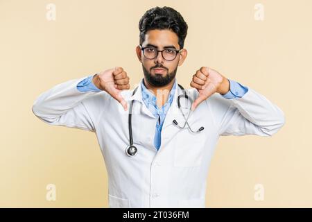 N'aime pas. Contrarié malheureux jeune médecin indien cardiologue homme montrant les pouces vers le bas signe geste, désapprobation, insatisfait, rétroaction négative. Homme de pharmacie apothicaire arabe isolé sur fond beige Banque D'Images