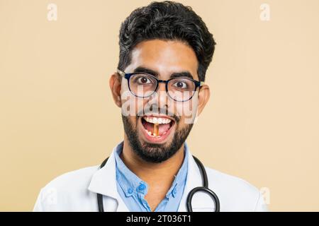 Homme de médecin indien souriant met de comprimé pilule vitamine D ou oméga-3 capsule jaune dans la bouche, recommande des médicaments de traitement de guérison d'immunisation. Les soins de santé. Pharmacien apothicaire sur fond beige Banque D'Images