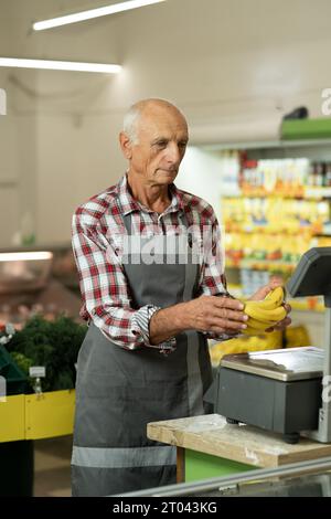 Vieux travailleur pesant des bananes sur des balances dans un magasin ou un supermarché. Concept de vente au détail Banque D'Images