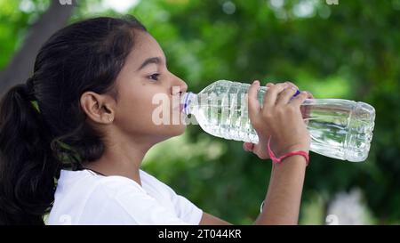 Heureuse fille indienne mignonne avec des cheveux noirs buvant de l'eau dans le parc, tenant la bouteille en plastique et montrant le pouce vers le haut. prise de vue en extérieur Banque D'Images