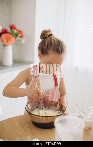 Une mignonne petite fille dans un tablier prépare de la pâte à crêpes dans la cuisine. Banque D'Images