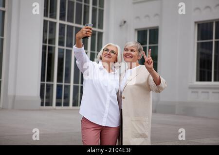 Belles femmes âgées prenant la photo selfie sur le téléphone portable à la ville, sourire sur fond de bâtiment blanc. Deux amies plus âgées en vacances. Banque D'Images