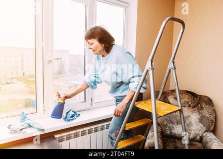 Fenêtre de nettoyage Happy Shy mature Woman avec Spray. Concept Seniors actif. Femme âgée nettoyage appartement. Femme au foyer. Femme caucasienne Moyen âge Banque D'Images