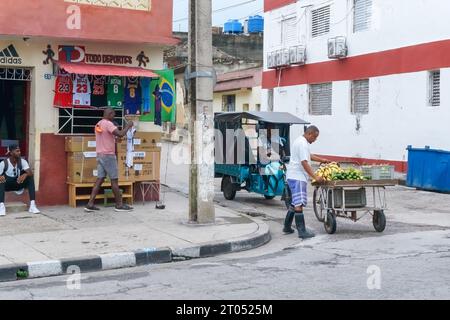 Un cubain vend des fruits et des légumes dans une charrette au coin de la rue. Une autre personne masculine met son kiosque de maison avec des articles de sport à vendre. Santa Clara, Banque D'Images