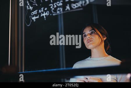 Pensive businesswoman contemple des idées derrière un mur de verre dans un bureau tard dans la nuit. Cet analyste de données créatif et entrepreneur travaille avec diligence, str Banque D'Images