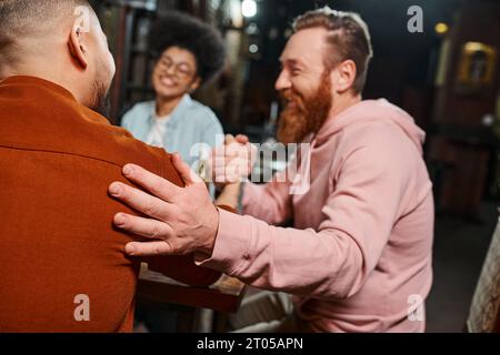 homme barbu heureux serrant la main avec un collègue dans un pub près d'une femme afro-américaine floue Banque D'Images