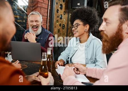homme barbu excité riant près d'un ordinateur portable et collègues de travail multiethniques avec des bouteilles de bière parlant dans le pub Banque D'Images
