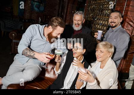 homme barbu tatoué claquant des lunettes avec des femmes gaies près d'amis multiethniques dans un bar à cocktails Banque D'Images