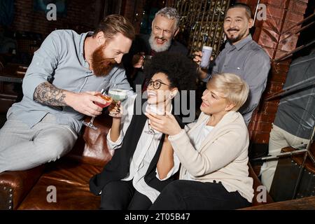 homme barbu tatoué claquant des lunettes avec des femmes multiethniques près de collègues souriants dans un bar à cocktails Banque D'Images