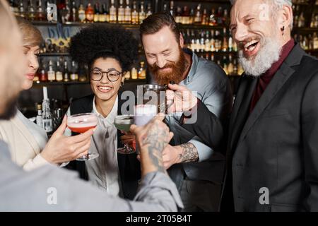 hommes barbus tatoués clinquant des lunettes avec des femmes multiethniques heureuses dans un bar à cocktails après le travail Banque D'Images