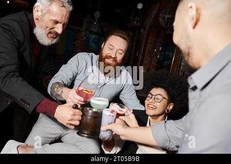 hommes barbus tatoués et femme afro-américaine clinquant des verres dans le bar à cocktails, après le plaisir du travail Banque D'Images