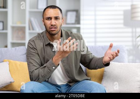 Portrait d'un jeune homme afro-américain inquiet assis à la maison sur le canapé, parlant sur un appel vidéo à la caméra, faisant des gestes avec ses mains. Banque D'Images