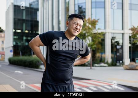 Jeune athlète asiatique masculin tenant son dos et ressentant de fortes douleurs. Debout dans une rue de ville et souffrant de douleur. Banque D'Images