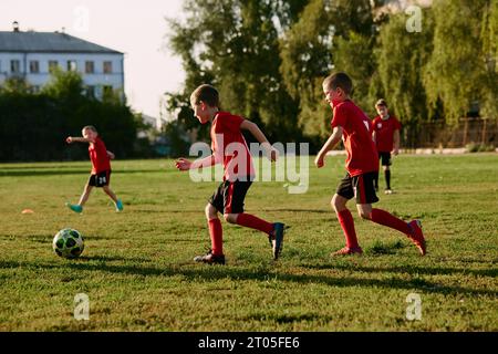 Photo en pied d'enfants, joueurs de football en uniforme de sport entraînement en mouvement avec balle avant le match sur le terrain de football sur la journée ensoleillée d'été. Banque D'Images