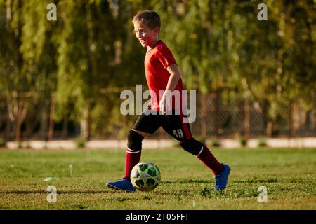 Portrait de vue latérale pleine longueur de l'enfant, joueur de football dans l'entraînement uniforme de sport, courir en mouvement avec le ballon avant le match sur le terrain de football. Banque D'Images