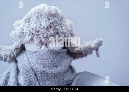 Un homme gelé tout empaqueté dans un chapeau de trappeurs de fourrure, une écharpe et une parka, couvert de neige et de gel essayant de rester au chaud un jour gris très froid d'hiver. Banque D'Images