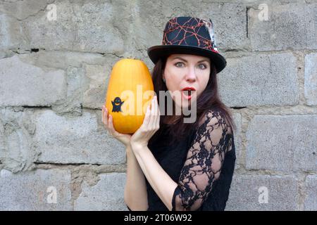 Gros plan portrait jeune femme en costume d'halloween exprimant la surprise. femme aux cheveux bruns en robe guipure noire et chapeau haut de forme tient la citrouille dans son han Banque D'Images