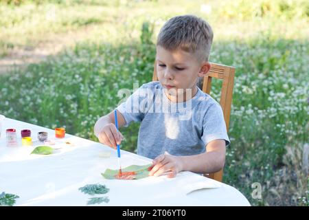 l'enfant peint des feuilles avec des peintures, dessine une image, fait des impressions de feuilles. La créativité des enfants dans la nature. Enseignement à distance. Extérieur. Été. Banque D'Images