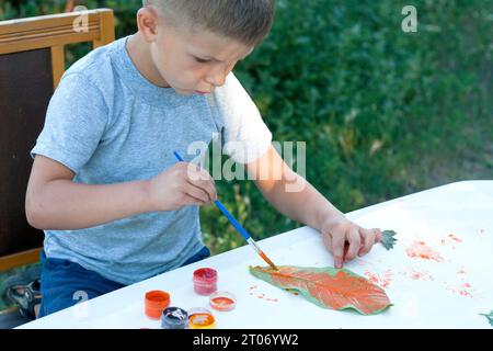 enfant peint les feuilles avec des peintures, dessine une image, faisant des impressions de feuilles. La créativité des enfants dans la nature. Extérieur. Été. Banque D'Images