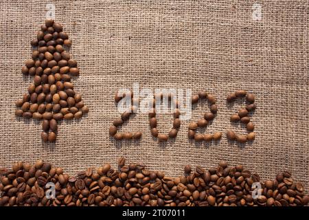 Joyeux Noël et carte de voeux du nouvel an. Numéros 2023 et arbre de Noël sont fabriqués à partir de grains de café sur tissu avec texture de toile de jute. Vue d'en haut. Banque D'Images