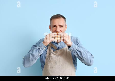 Homme mature mangeant un hamburger savoureux sur fond bleu Banque D'Images