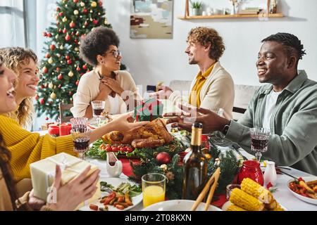 Grande famille multiculturelle assise à la table de fête échangeant des cadeaux avec sapin de Noël en toile de fond Banque D'Images