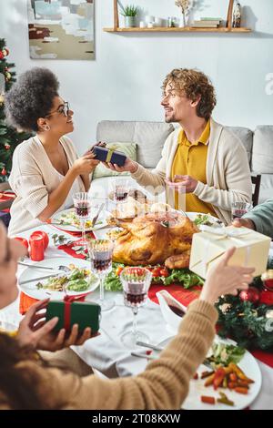 Jeunes joyeux membres de la famille multiethnique assis à la table de vacances et échangeant des cadeaux, Noël Banque D'Images