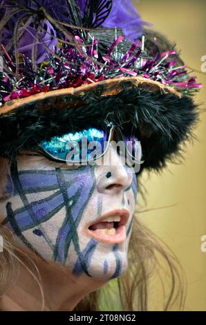 femme dans le chapeau de fantaisie et lunettes de soleil avec peinture de visage sur la bouche ouverte Banque D'Images