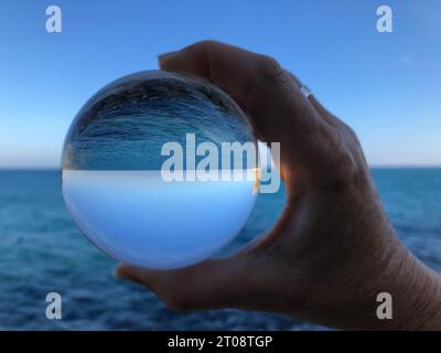 La main de femme tenant une boule de cristal, à la recherche jusqu'à l'océan et le ciel. La photographie créative, crystal ball réfraction Banque D'Images