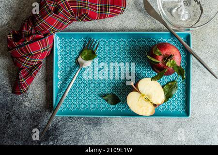 Vue de dessus de pomme mûrie rouge fraîche entière avec des feuilles vertes et demi-pièce coupée placée sur le plateau bleu près du verre à eau de serviette à carreaux sur la surface grise i Banque D'Images