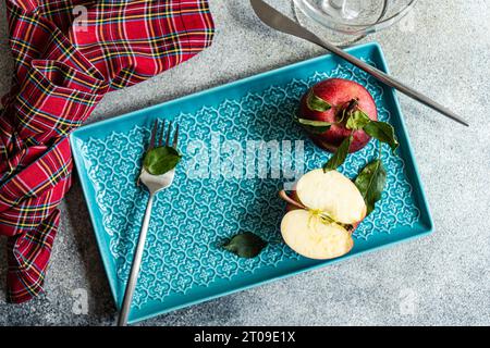 Vue de dessus de pomme mûrie rouge fraîche entière avec des feuilles vertes et demi-pièce coupée placée sur le plateau bleu près du verre à eau de serviette à carreaux sur la surface grise i Banque D'Images