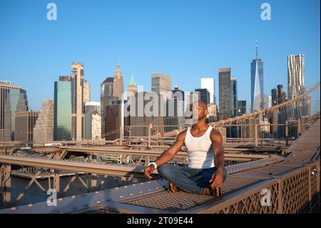 Homme afro-américain assis avec les jambes croisées tout en méditant à l'infrastructure du pont de Brooklyn à suspension métallique avec les yeux fermés dans la ville de jour Banque D'Images